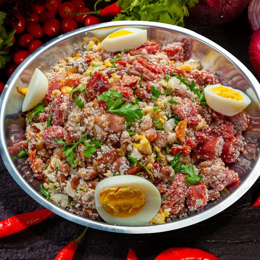 foto de uma tigela com feijão tropeiro e ovos cozidos por cima. Ao fundo, pimentas, tomates e cebolas picadas.