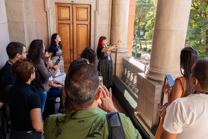 Grupo de nove pessoas observando os jardins do  MM Gerdau - Museu das Minas e do Metal, sendo uma delas a mediadora da visita, explicando detalhes ao grupo, que é composto por homens e mulheres.